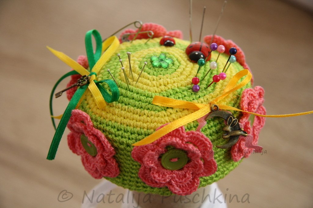 Rundes gehäkeltes Nadelkissen in Gelb und Grün mit rosa Blumen, bunten Bändern, Anhängern und Stecknadeln