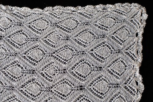 Weißes gestricktes Lacetuch mit Rautenmuster und gewellter Spitzenkante auf schwarzem Hintergrund.