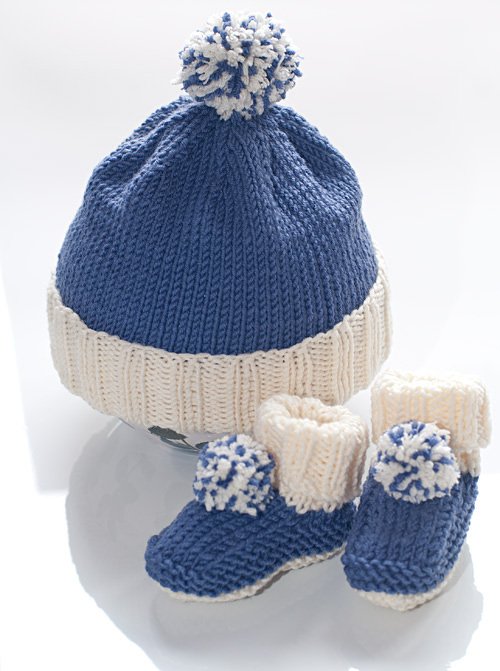 Blau-weiße gestrickte Babymütze mit Umschlag und Bommel neben passenden blauen Babyschühchen mit cremefarbenen Bündchen und Bommeln vor hellem Hintergrund.