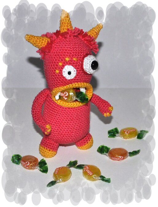 Gehäkeltes rotes Monster mit gelben Hörnern, großen Augen und Bonbon im Mund; verpackte Bonbons auf dem Boden