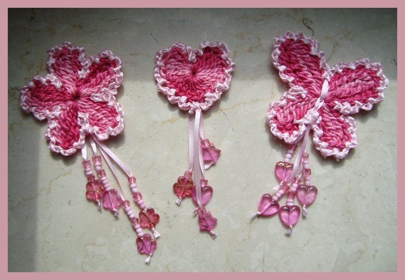 Drei rosa gehäkelte Applikationen: zwei floral-/schmetterlingsförmige Motive und ein Herz, verziert mit Perlen und schmalen Bändern.