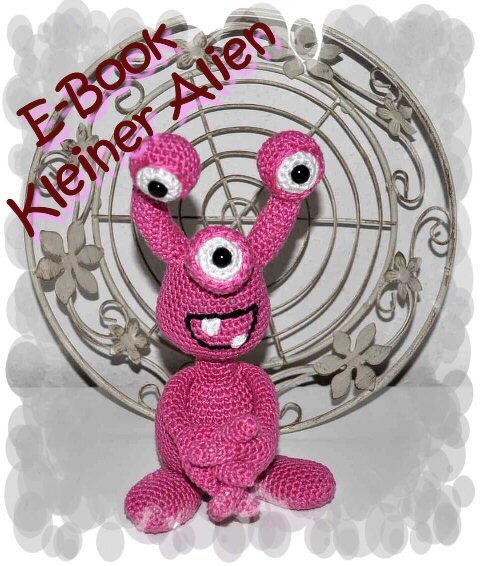 Rosa gehäkeltes Amigurumi mit drei Augen vor dekorativem Hintergrund; eingeblendeter Text 'E-Book Kleiner Alien'.