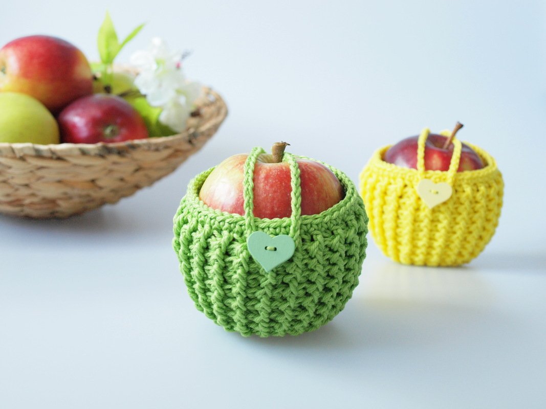 Zwei Äpfel in gehäkelten Täschchen (grün und gelb) mit herzförmigen Knöpfen; im Hintergrund ein Korb mit Äpfeln.