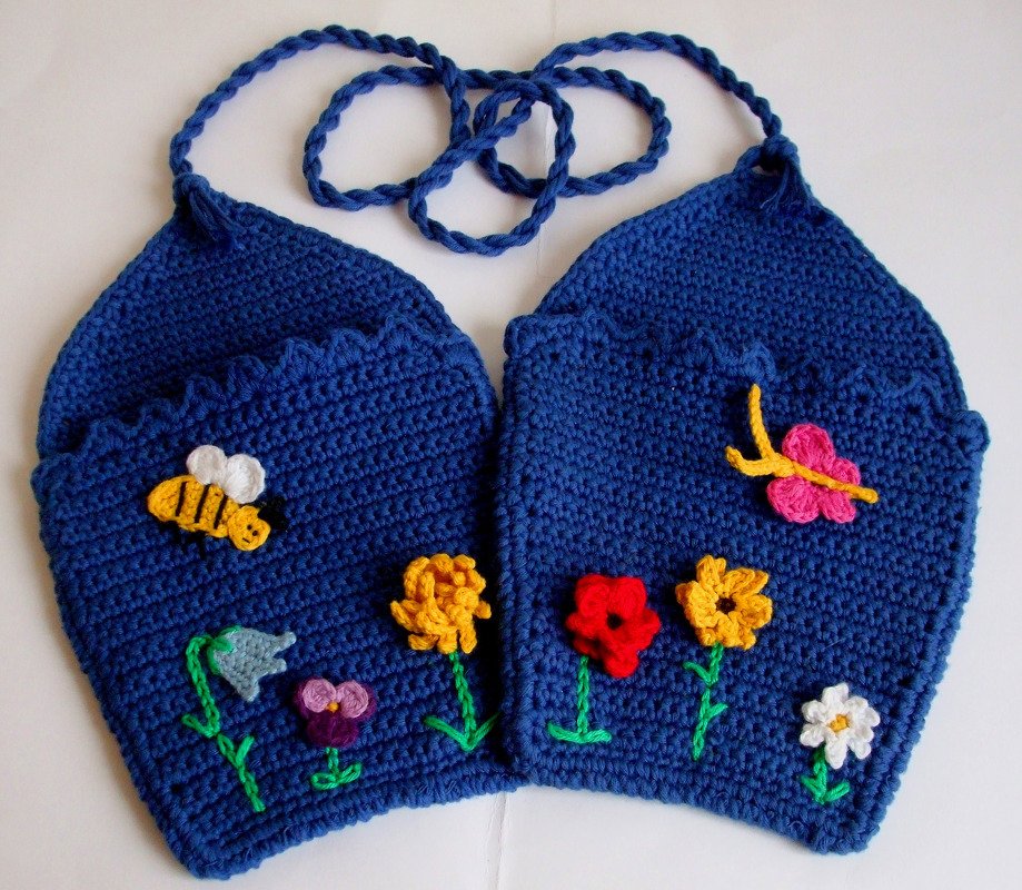Zwei blaue gehäkelte Topflappen mit applizierten Blumen, einer Biene und einem Schmetterling, verbunden durch eine geflochtene Kordel.