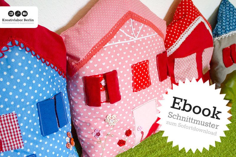 Drei dekorative Hauskissen in Rosa, Blau und Rot mit applizierten Fenstern, Türen und Blumenmotiven.