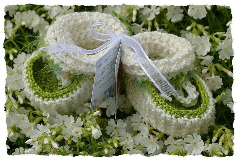 Gehäkelte Babyschuhe cremeweiß mit grünem Rand und weißer Schleife auf weißen Blüten