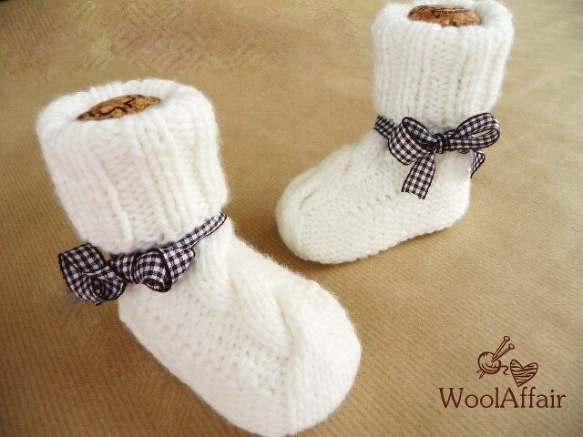 Paar weißer, handgestrickter Babyschuhe mit umgeschlagenem Schaft und kariertem Schleifenband auf beigem Hintergrund