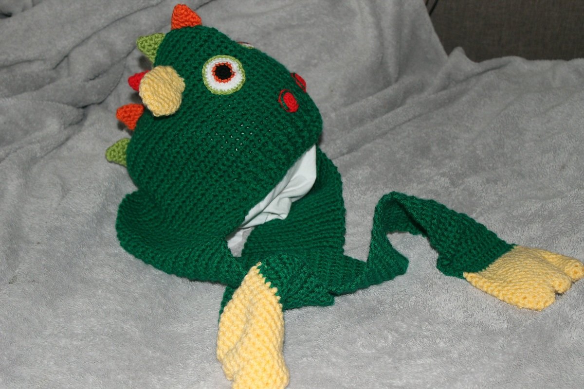 Grüne gehäkelte Babymütze im Drachen-Design mit Augen, bunten Stacheln und langen Ohrenklappen mit gelben Enden