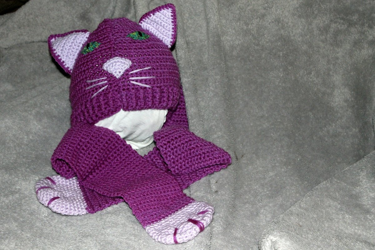 Lila gehäkelte Katzenmütze mit helllila Innenohren, grüner Bestickung für die Augen, gestickter Nase und langen Ohrenklappen mit Pfötchenenden auf grauem Hintergrund.