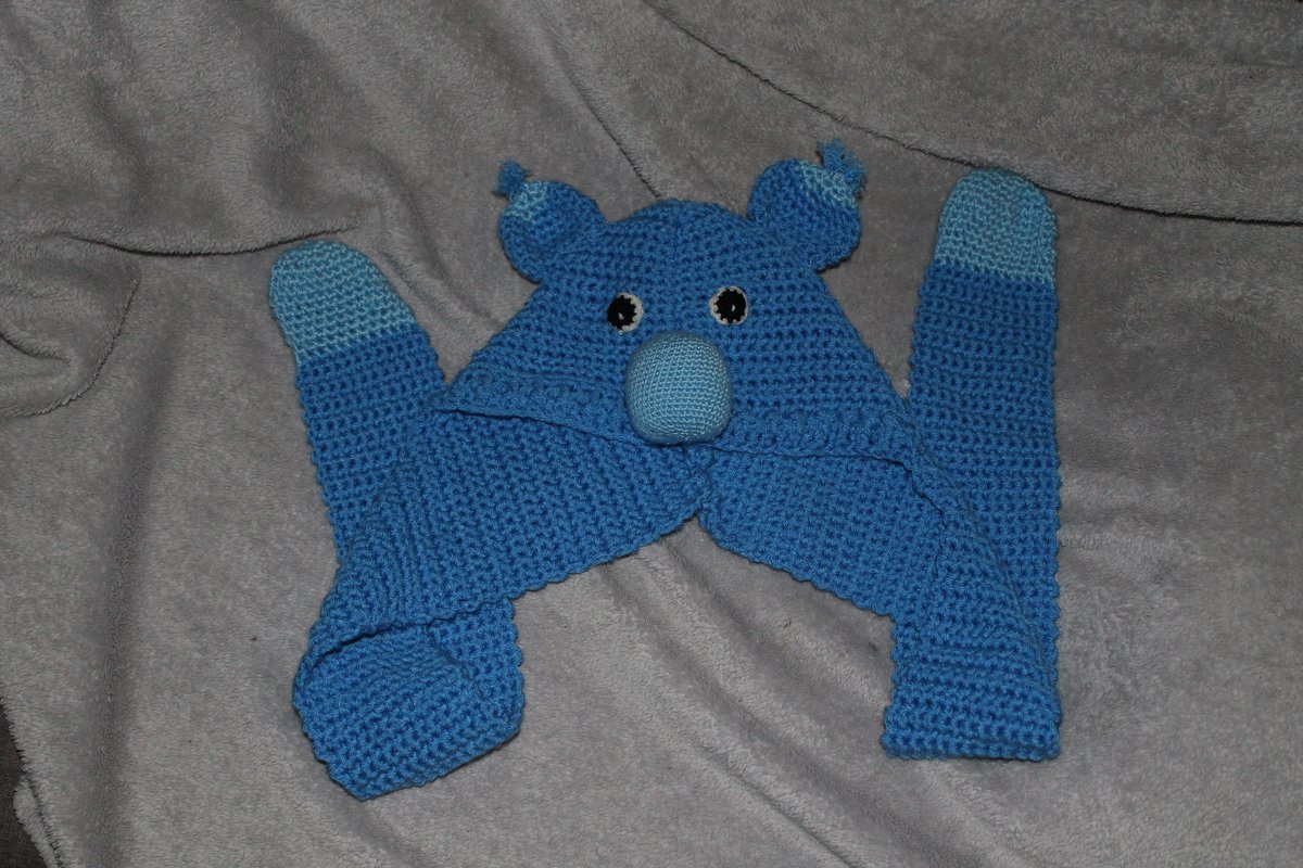 Blaue gehäkelte Kindermütze mit Tiergesicht, Ohren und langen Schalenden auf grauem Hintergrund.