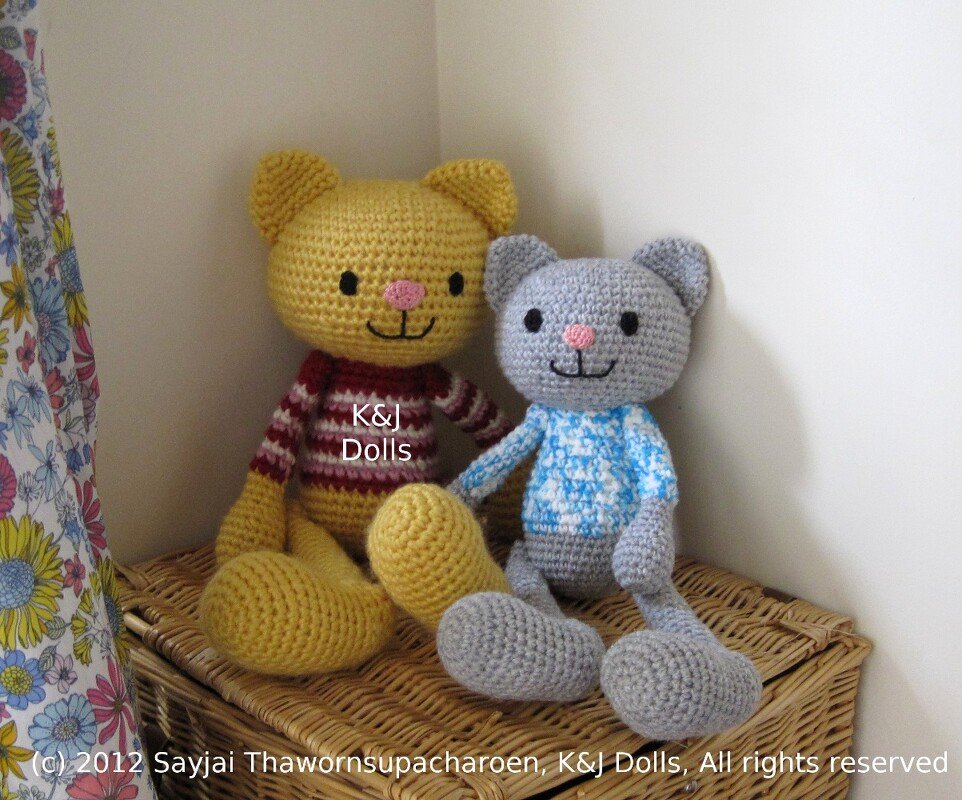 Two crocheted amigurumi cat dolls
— yellow with red-striped sweater and grey with blue patterned sweater
— seated on a woven basket