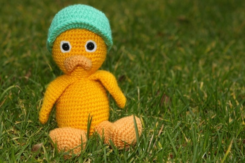 Handgehäkelte gelbe Amigurumi-Ente mit mintgrüner Mütze sitzt im Gras.