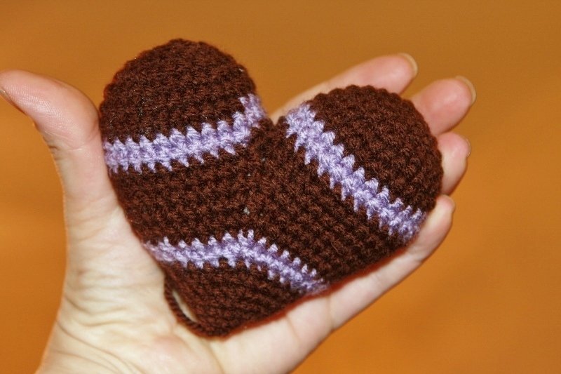 Hand hält ein braunes gehäkeltes Amigurumi-Herz mit lila Streifen