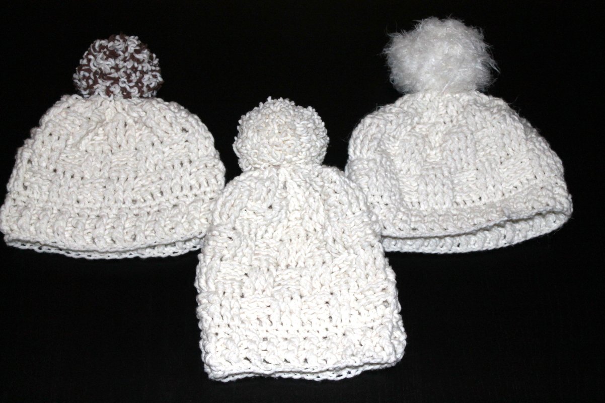 Drei weiße gehäkelte Mützen mit Bommeln auf schwarzem Hintergrund; eine Mütze hat einen braun-weißen Bommel.