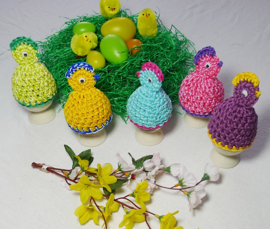Sechs bunte gehäkelte Küken-Eierwärmer auf Eierbechern vor grünem Ostergras und bunten Deko-Eiern