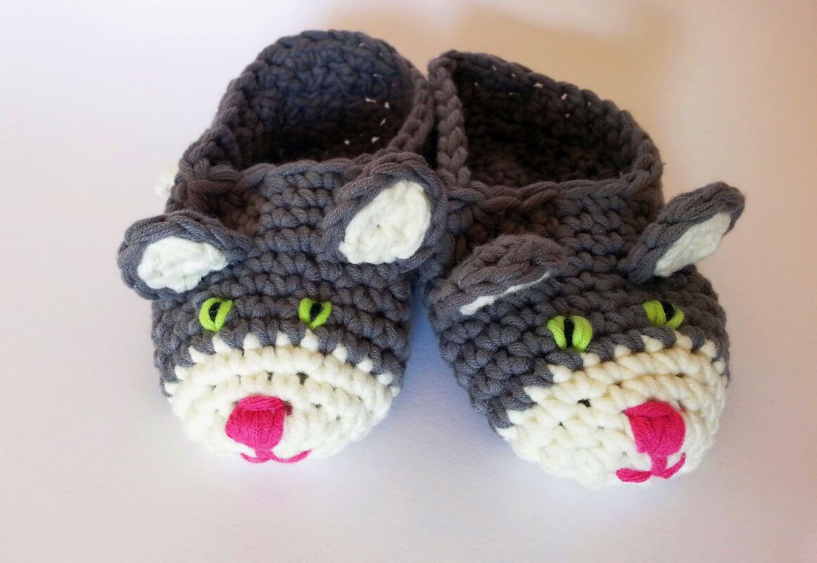 Nahaufnahme gehäkelter grauer und weißer Kinderpantoffeln mit Katzenohren, grünen Augen und rosa Schnauze