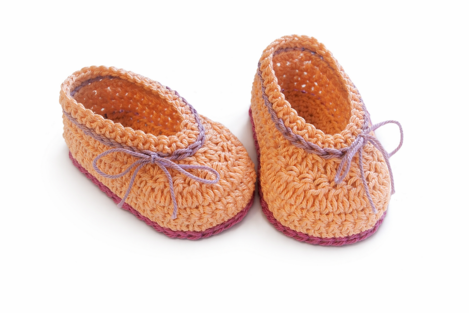 Zwei gehäkelte Babyschuhe in Pfirsich mit lila Sohle und lila Bindeband auf weißem Hintergrund.
