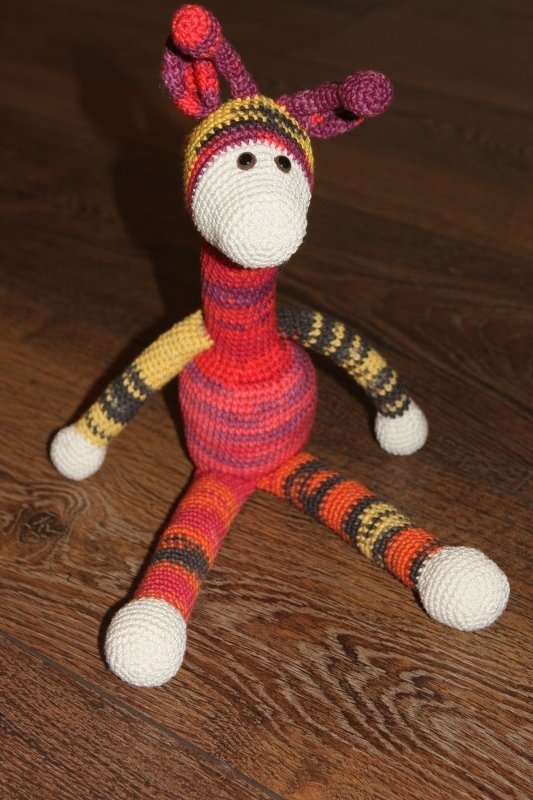 Gehäkeltes Amigurumi-Tier mit langem Hals und gestreiften Beinen, sitzend auf Holzfußboden
