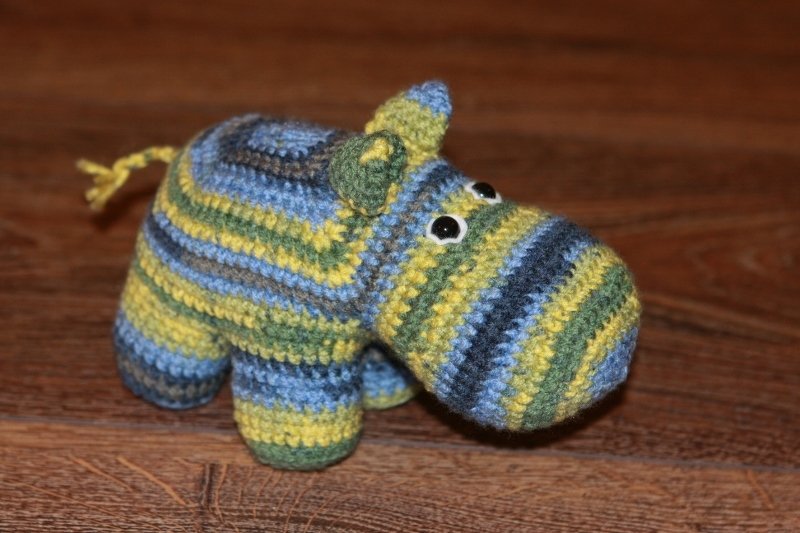 Gehäkeltes Amigurumi-Nilpferd mit blau-grün-gelben Streifen und schwarzen Sicherheitsaugen auf Holzoberfläche.