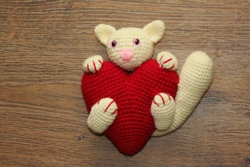 Gehäkelte Katze in Cremefarbe hält rotes Herz, Nahaufnahme