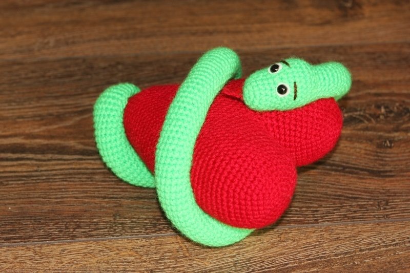 Grüne Amigurumi‑Schlange mit schwarzen Augen umschlingt ein rotes Amigurumi‑Herz auf Holz.