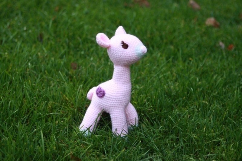 Rosa gehäkeltes Rehkitz (Amigurumi) mit dunklen Augen und lila Akzent, stehend im grünen Gras.