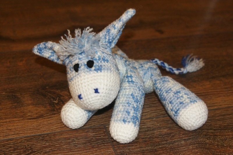 Gehäkeltes blau-weißes Amigurumi-Eselchen mit Fransenmähne und geflochtenem Schwanz auf Holzfußboden