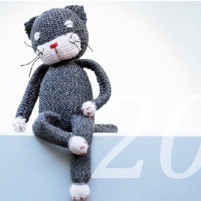 Gestrickte graue Amigurumi-Katze mit weißer Schnauze und roter Nase, sitzend auf einer hellen Fläche.