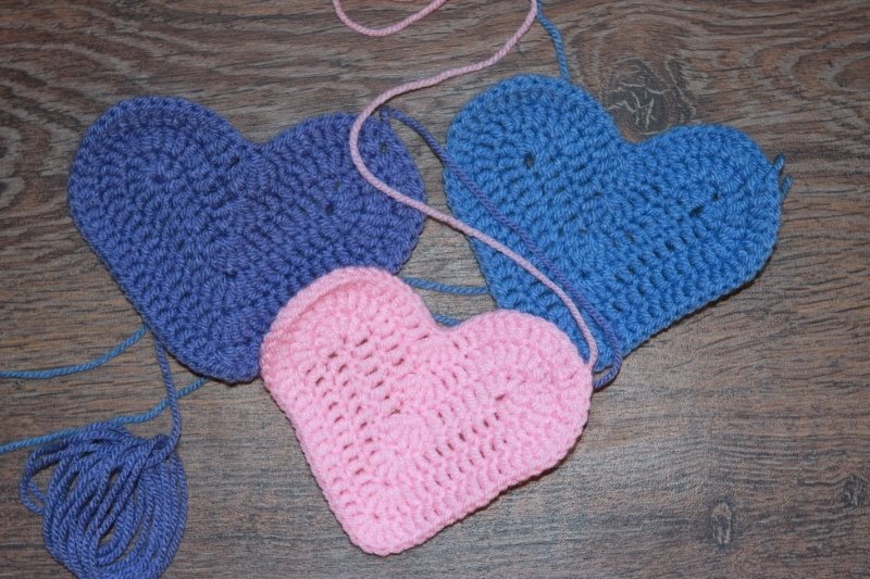Drei gehäkelte Herz-Applikationen in Blau und Rosa auf Holz mit sichtbaren Garnfäden