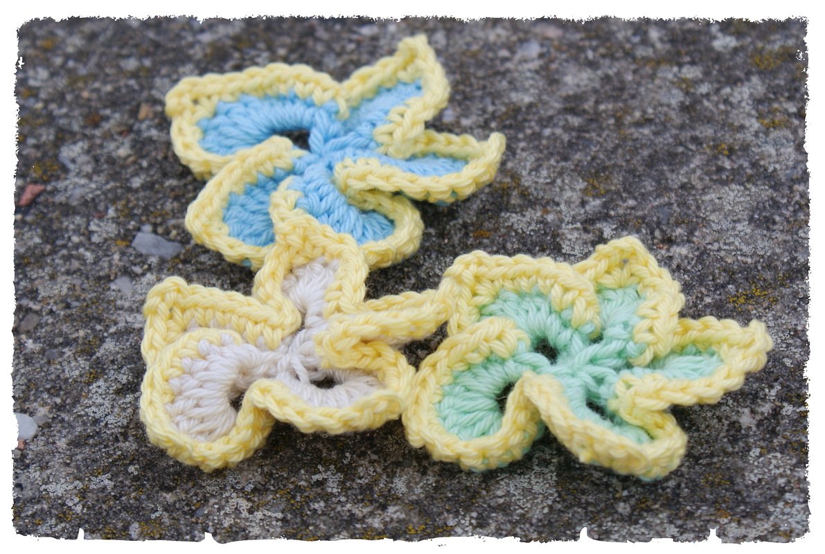 Drei pastellfarbene, fünfblättrige gehäkelte Blumen mit gelber Umrandung auf Stein