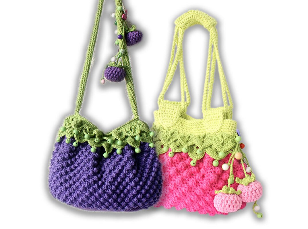 Two crocheted shoulder bags: purple with green scalloped trim and purple berry charms; pink with green scalloped trim, yellow straps, and pink berry charms.