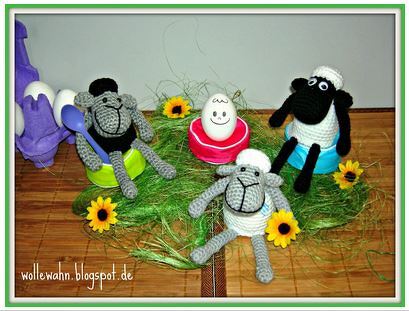 Drei gehäkelte Schafe als Osterdekoration mit einem lachenden Ei im roten Eierbecher, gelben Blumen und künstlichem Gras auf einem Holztisch.