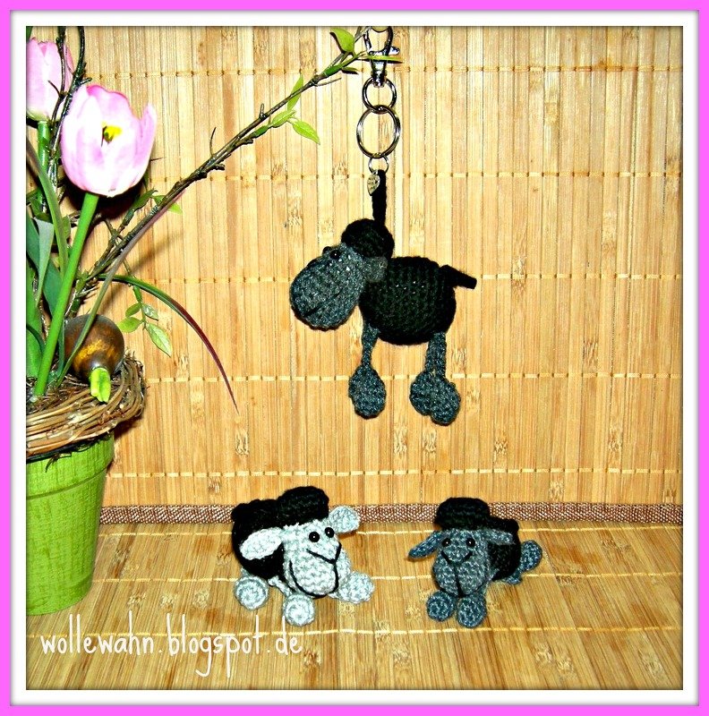 Drei gehäkelte Schäfchen, eines als Schlüsselanhänger hängend, zwei stehende Figuren auf Holz vor Bambusmatte und Pflanze.