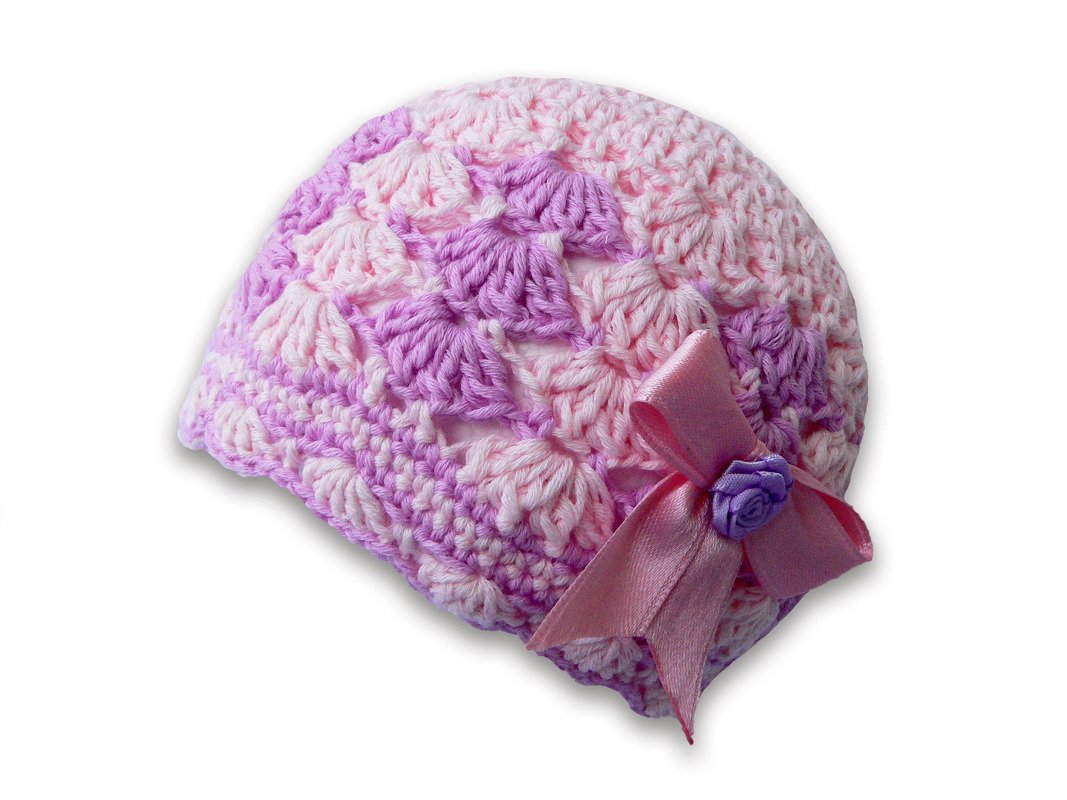 Nahaufnahme einer gehäkelten Babymütze in Rosa und Lila mit Muschelmuster, rosa Satinschleife und kleinem lila Häkelblümchen