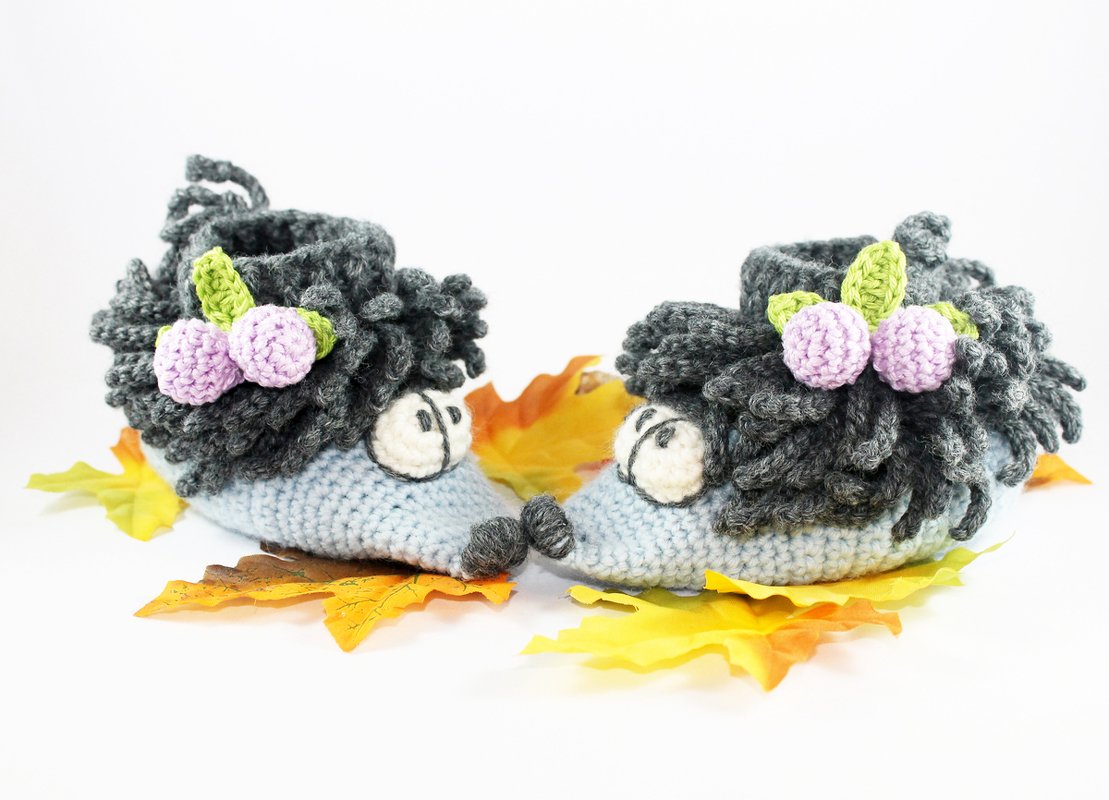 Paar gehäkelter Igel-Babyschuhe in Grau und Hellblau mit lila Beeren auf orange-gelben Herbstblättern.