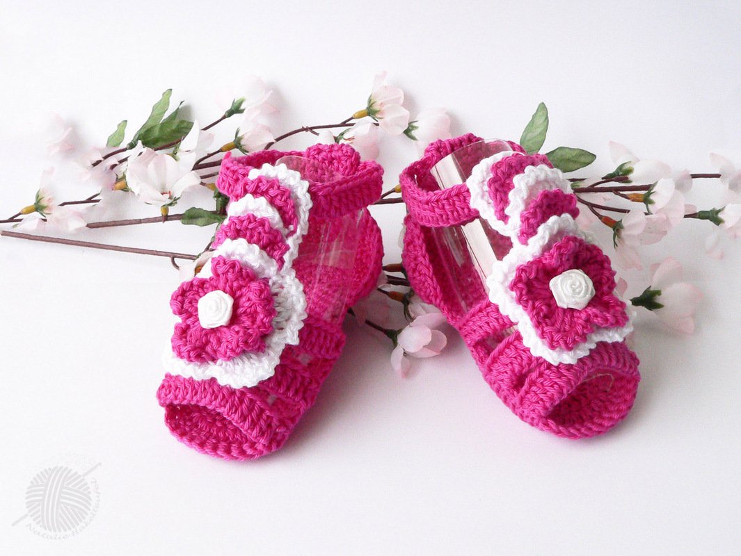 Pink-weiße gehäkelte Babysandalen mit Blüten, arrangiert neben rosa Blütenzweigen.