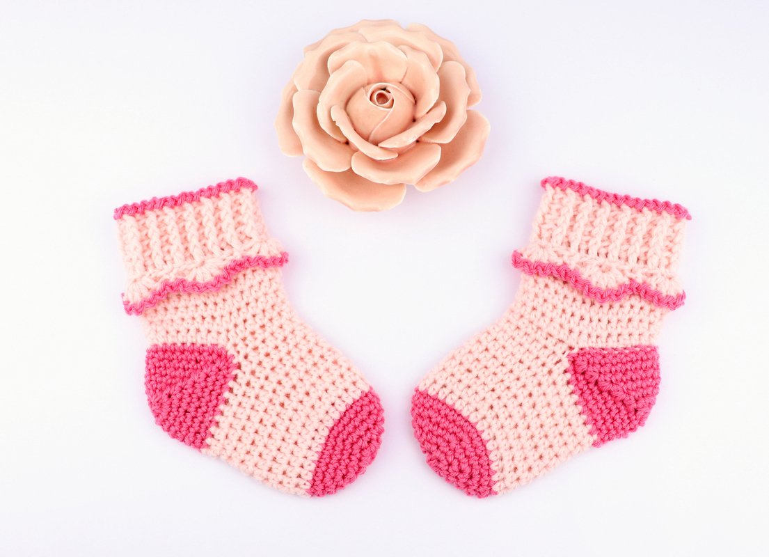 Paar gehäkelter Babysocken hellrosa mit dunkelpinkem Zehen- und Fersenbereich neben rosafarbener Blüte