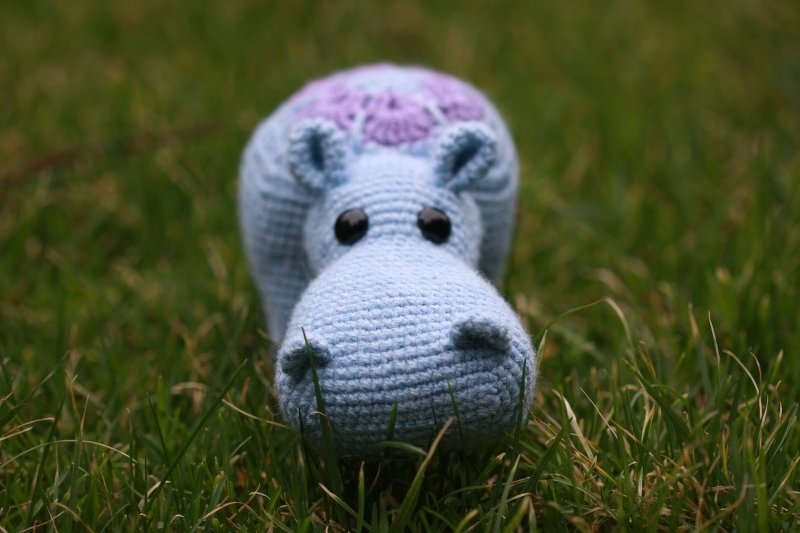 Hellblaues Amigurumi-Nilpferd mit lila Muster und schwarzen Augen auf Gras.