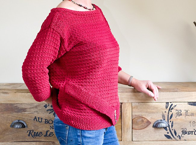 Roter, strukturiert gestrickter Damenpullover mit U‑Boot‑Ausschnitt und seitlichen Zierknöpfen, getragen zu blauen Jeans.