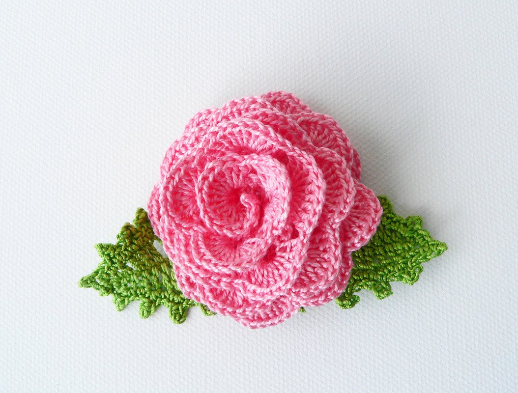 Pink crocheted rose with two green crocheted leaves on a white textured background