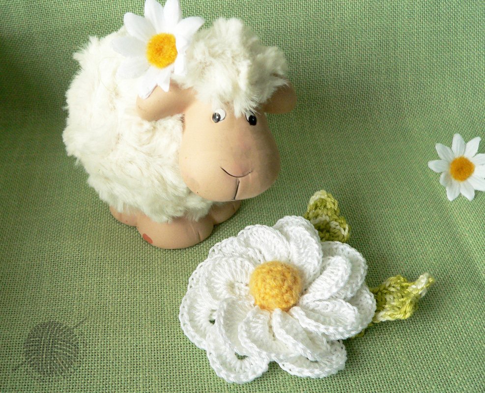 Weiße gehäkelte Margerite-Applikation mit gelbem Zentrum und zwei grünen Blättern vor grünem Stoff, neben einem Stoffschaf