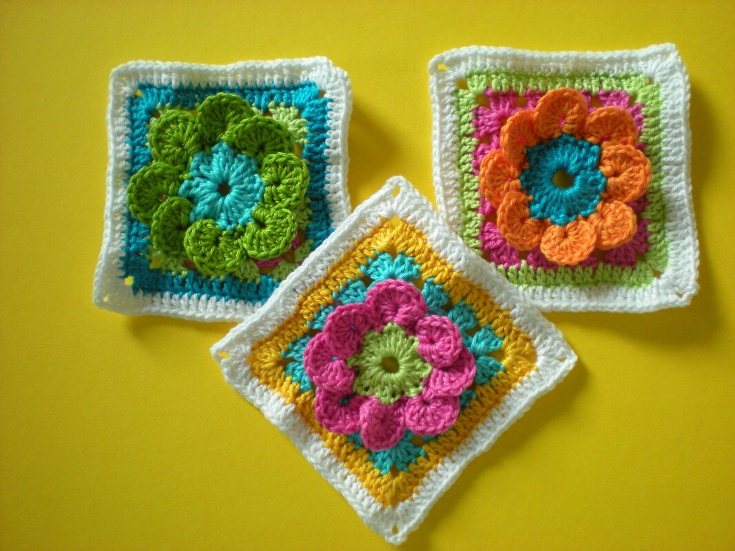 Drei gehäkelte Granny Squares mit erhabenen Blumenapplikationen in Pink, Orange und Grün auf gelbem Untergrund