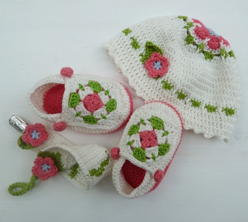 Gehäkeltes Babyset in Cremeweiß mit Babymütze, Babyschühchen und Band mit Clip, dekoriert mit rosa Häkelblumen und grünen Blättern.