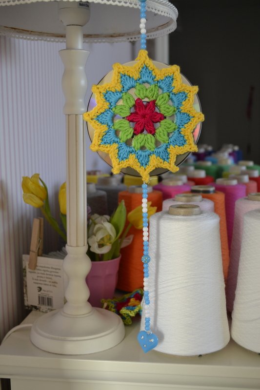 Gehäkeltes Mandala mit Perlenkette und blauem Herz vor Garnspulen