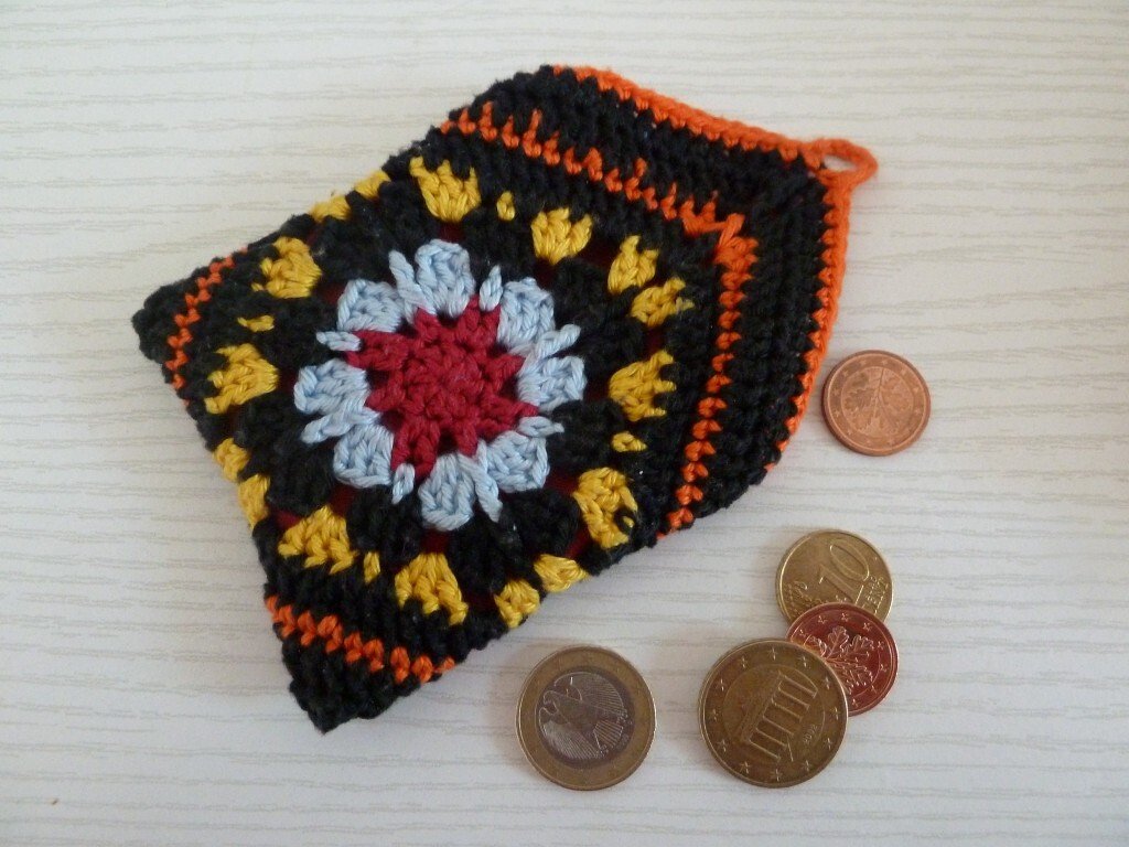 Gehäkelte schwarze Granny-Square-Börse mit bunten Akzenten, daneben Euro-Münzen