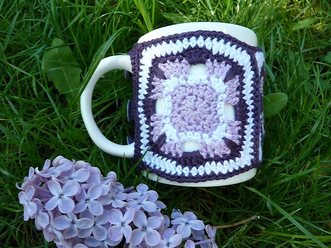 Weiße Tasse mit gehäkelter Granny-Square Hülle in Flieder- und Weißtönen auf Gras mit lila Blüten