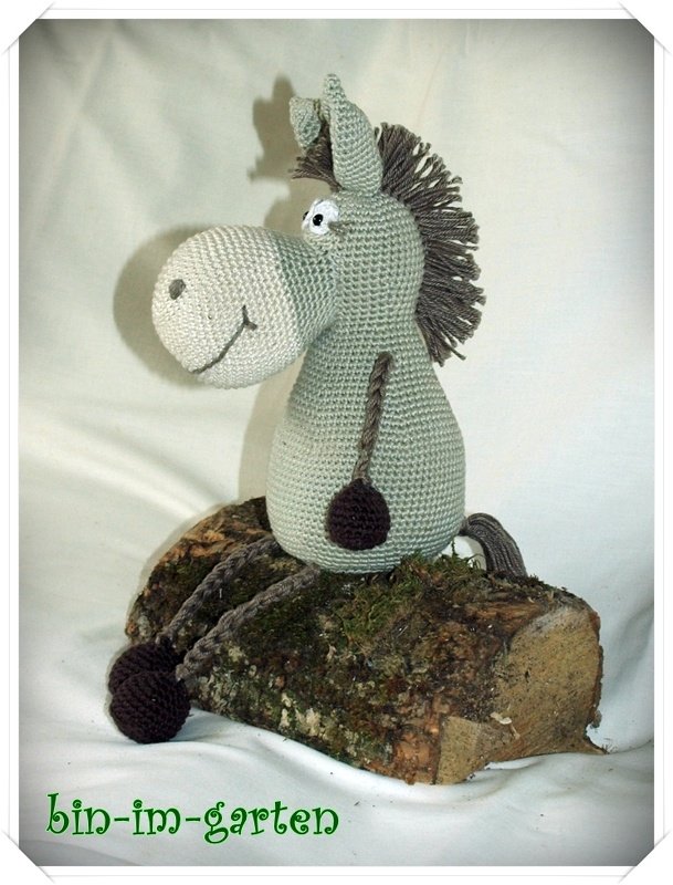 Pastellgrüner gehäkelter Amigurumi-Esel mit brauner Fransenmähne sitzend auf einem Holzstück