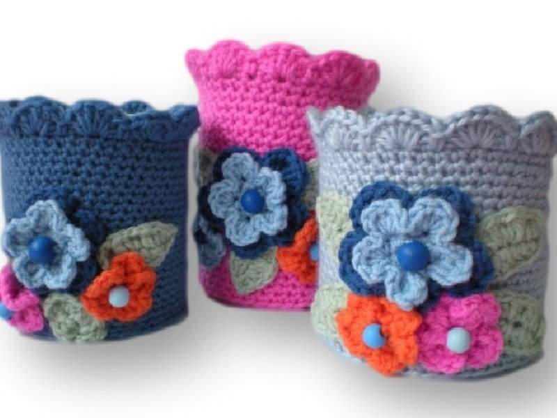 Drei gehäkelte Utensilos in Blau, Rosa und Hellgrau mit Blumenapplikationen