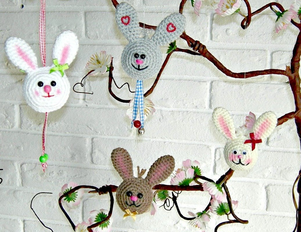 Mehrere gehäkelte Hasenköpfe in Weiß, Grau und Braun mit Schleifen und Verzierungen hängen an einem blühenden Zweig vor einer weißen Ziegelwand.