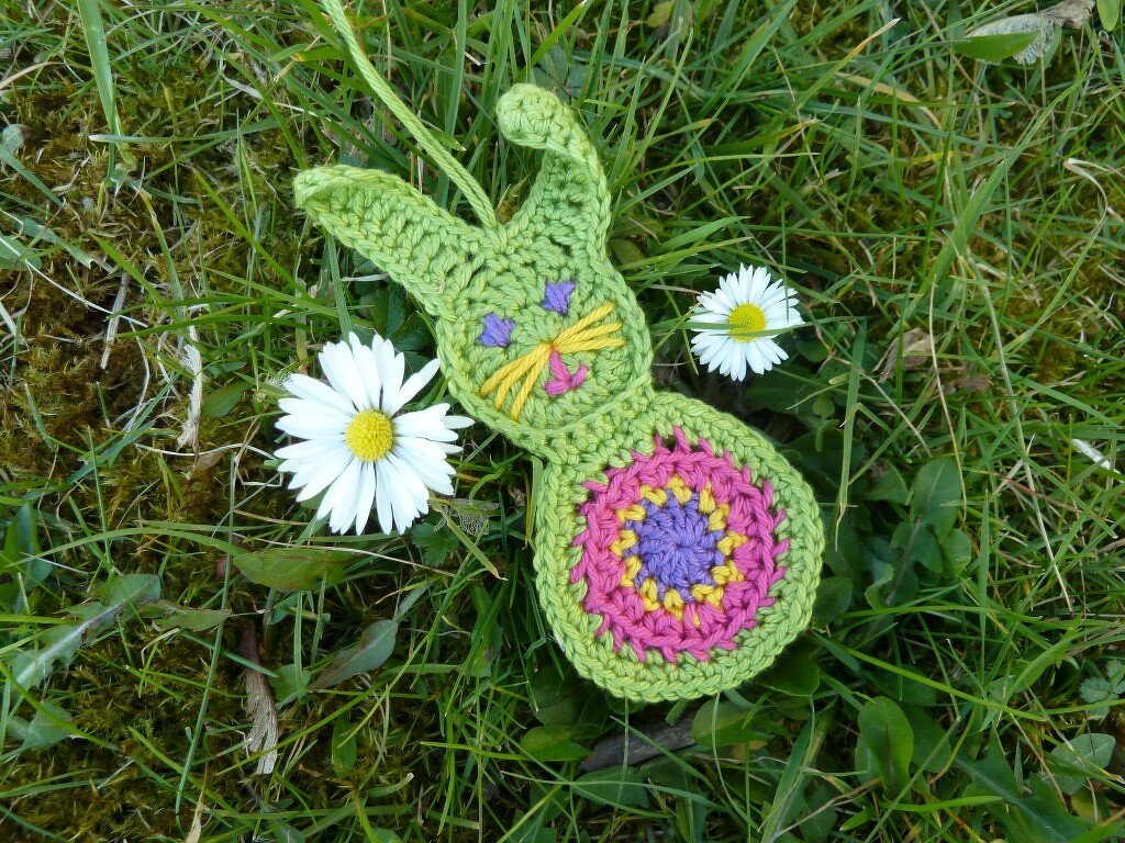Grüne gehäkelte Hasen-Applikation mit buntem Rundmuster auf Gras neben Gänseblümchen